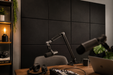 A home studio setup featuring a black acoustic foam panel wall, a wooden desk with a laptop, headphones, and two microphones on adjustable arms, with a shelving unit on the left.